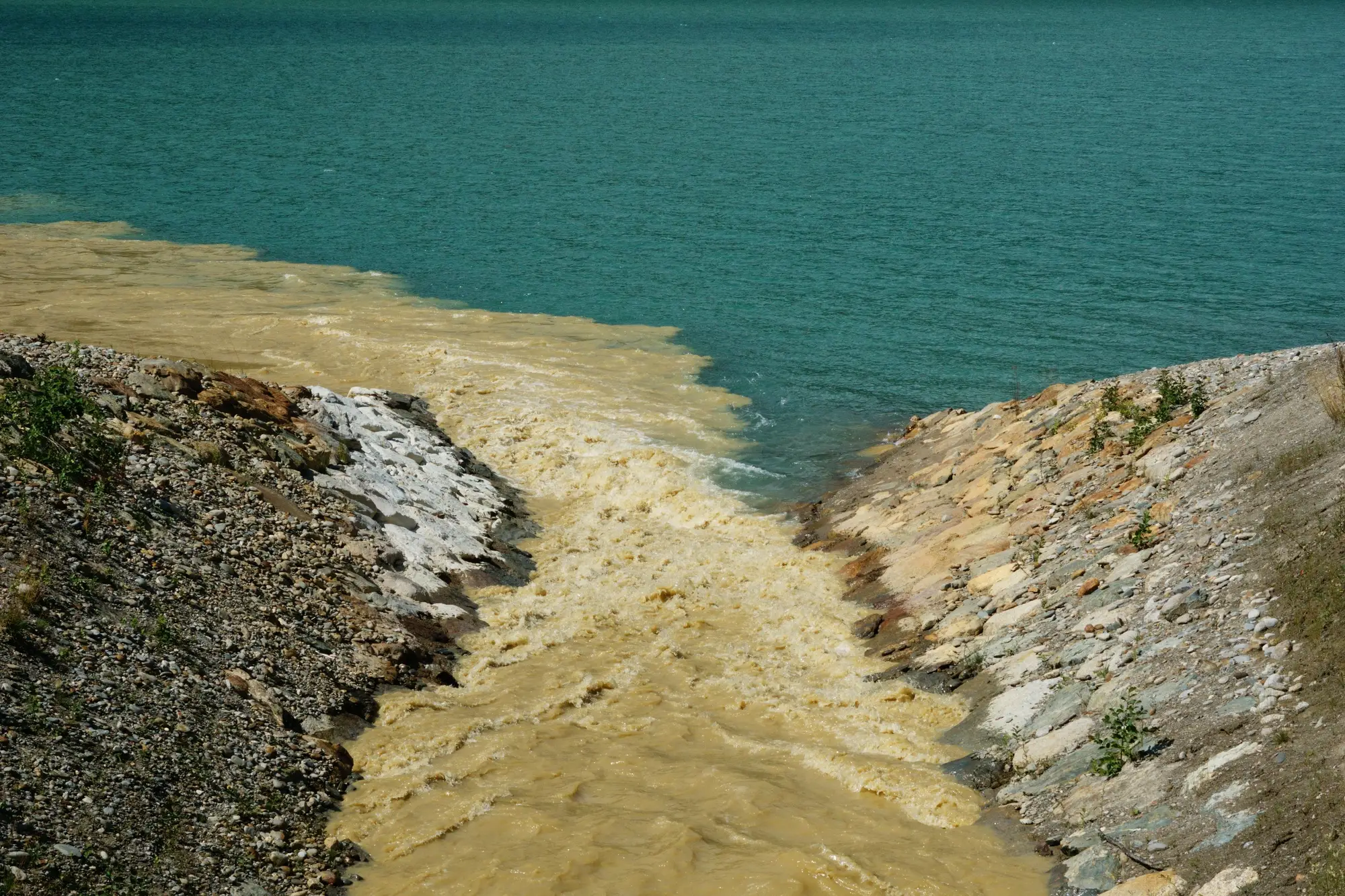 Silt-laden river water spilling out between stony banks into a clear green lake